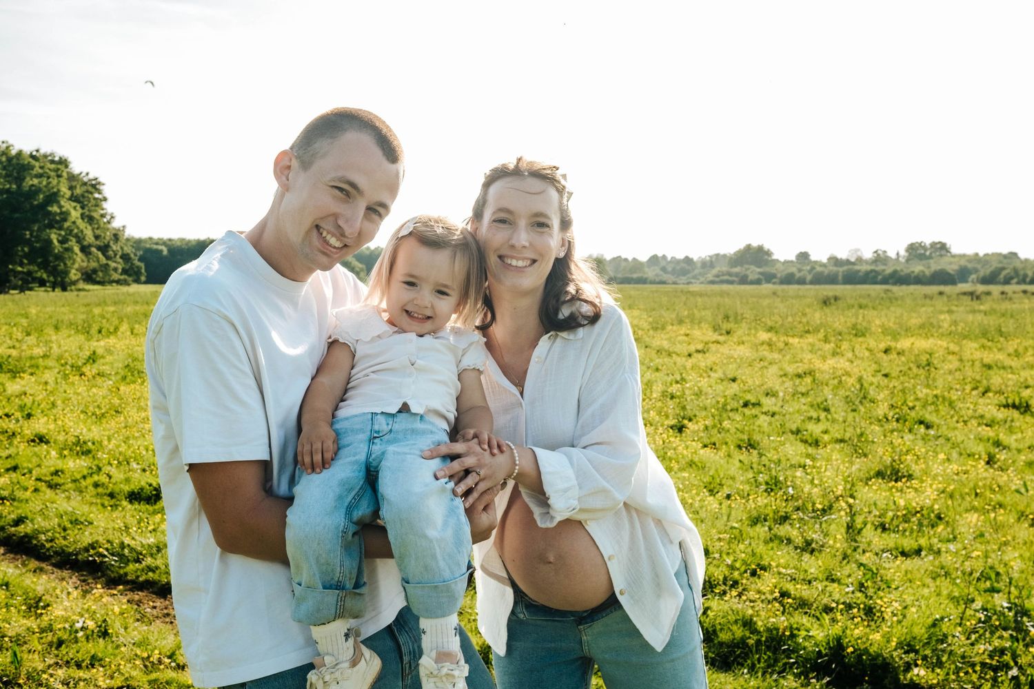 Happy family with toddler posing in sunny green field, pregnant mother and father smiling outdoors.