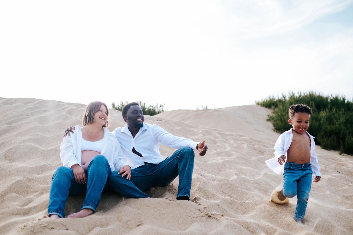 Interracial family relaxing on sandy beach dunes, parents sitting while toddler runs playfully nearby.