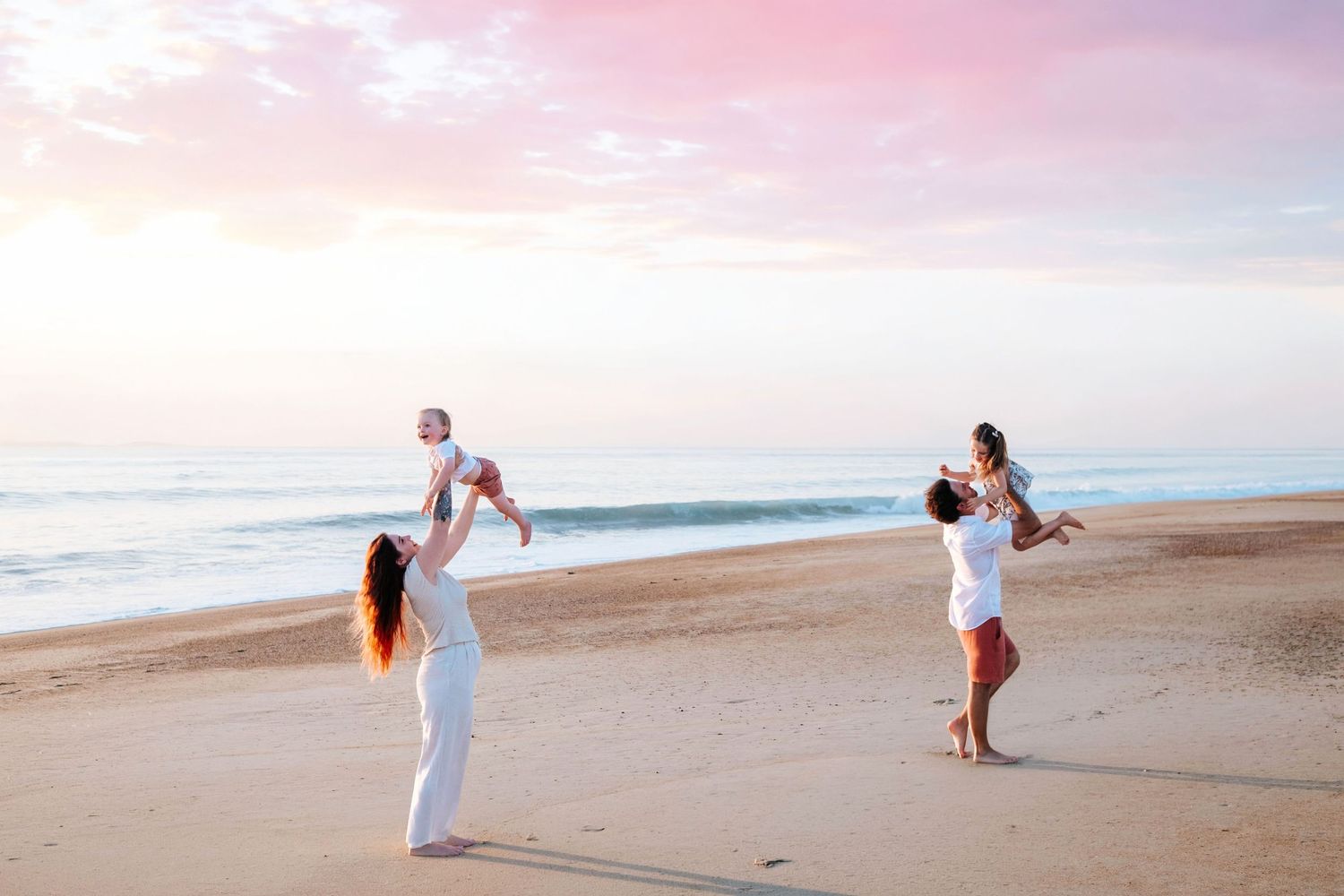 Family in white outfits playing on a sandy beach at sunset, parents lifting babies in the air near ocean waves.