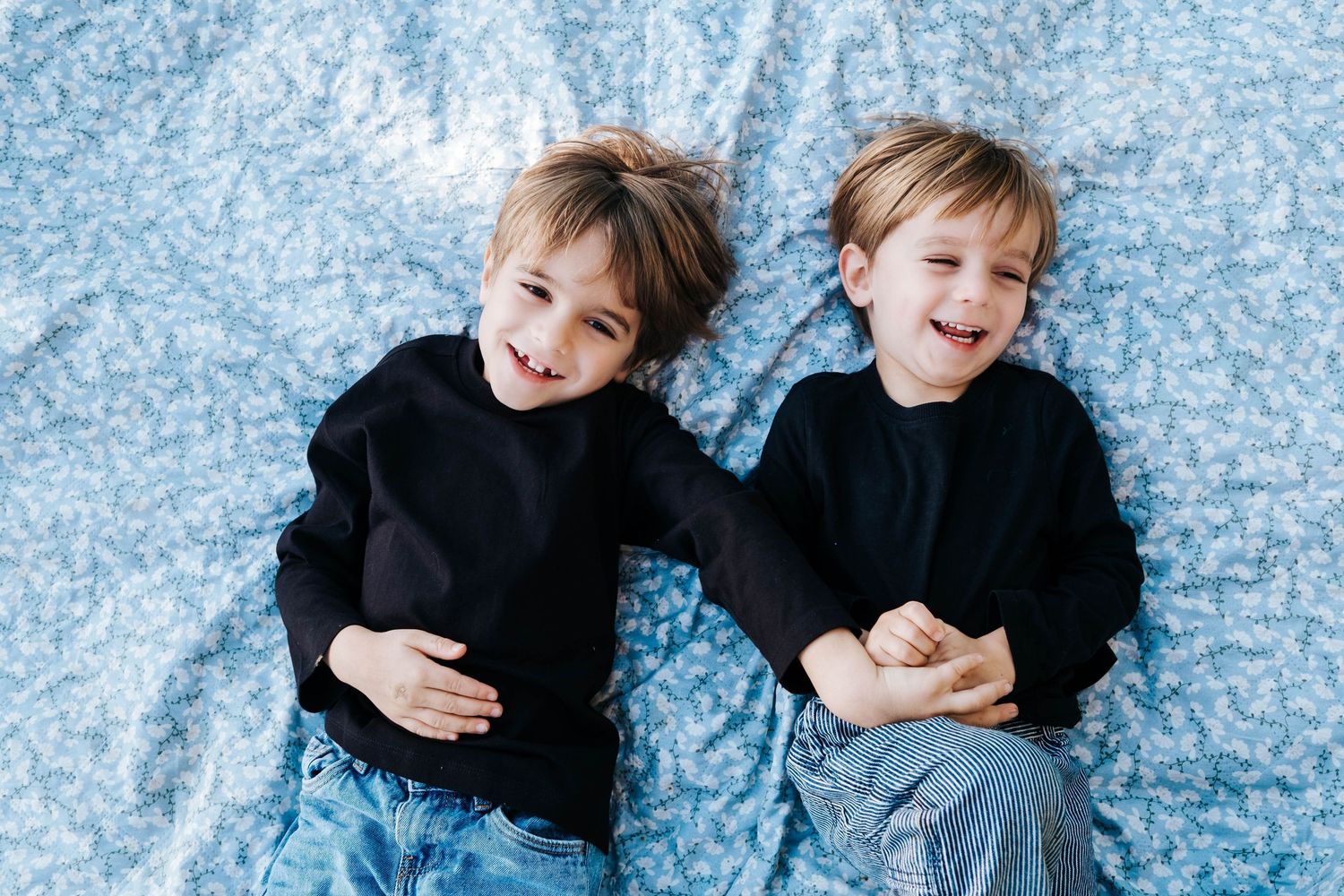 Two smiling boys in black sweaters lying on a blue floral blanket, laughing and hugging together.