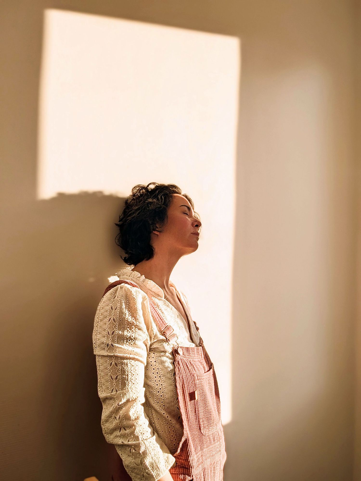 A series of four artistic portraits showing a person in a white dress against a soft, warm-toned wall with dramatic lighting.