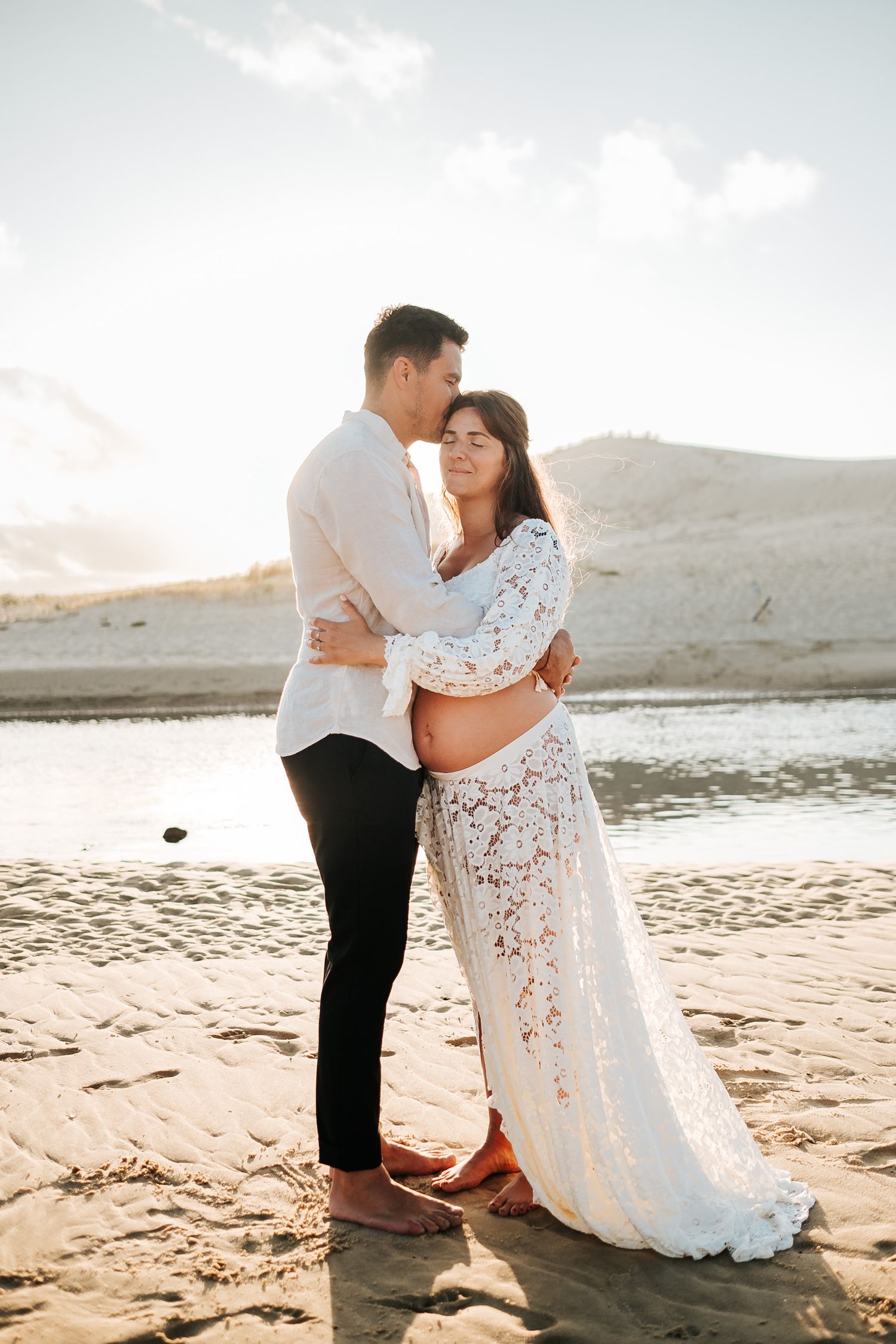 A couple shares a romantic moment on a sunlit beach in Hawaii, with the bride wearing a flowing white lace dress.