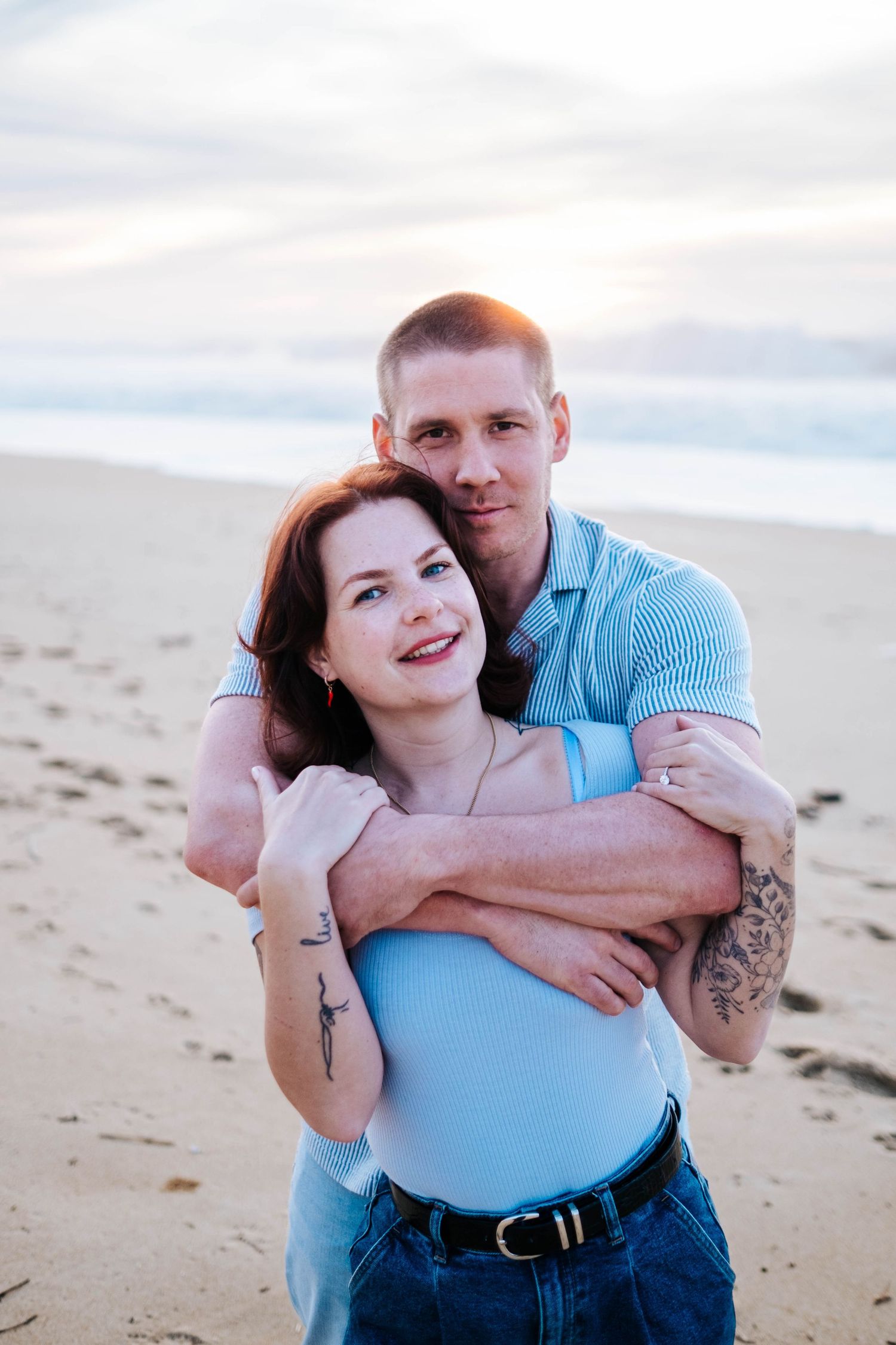 A couple embraces on a sandy beach during sunset wearing blue and denim clothing.