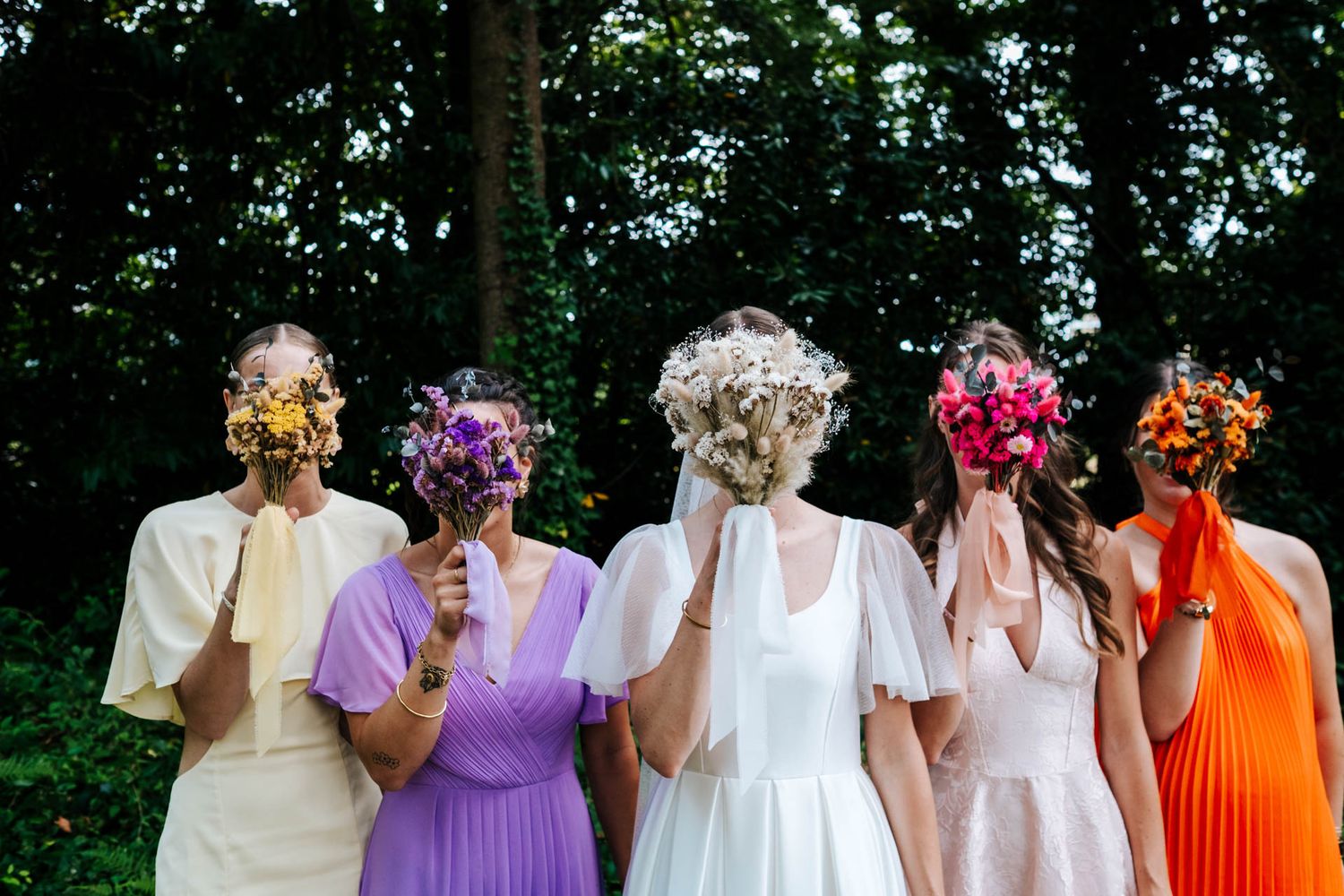 Five people in colorful dresses stand in a row wearing vibrant floral masks against a forest backdrop.