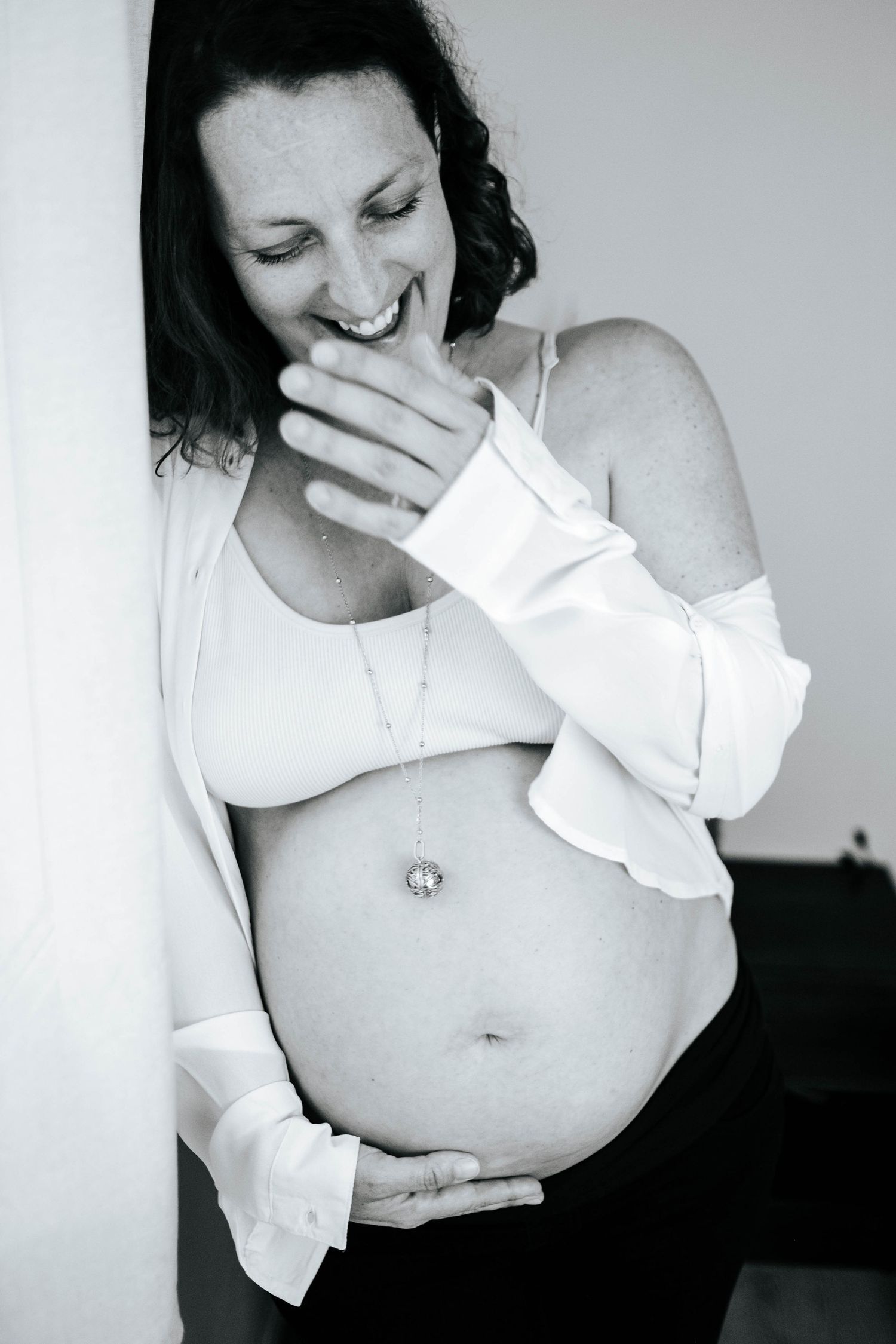 Black and white photo of a smiling pregnant woman cradling her baby bump, wearing an open white shirt and necklace.