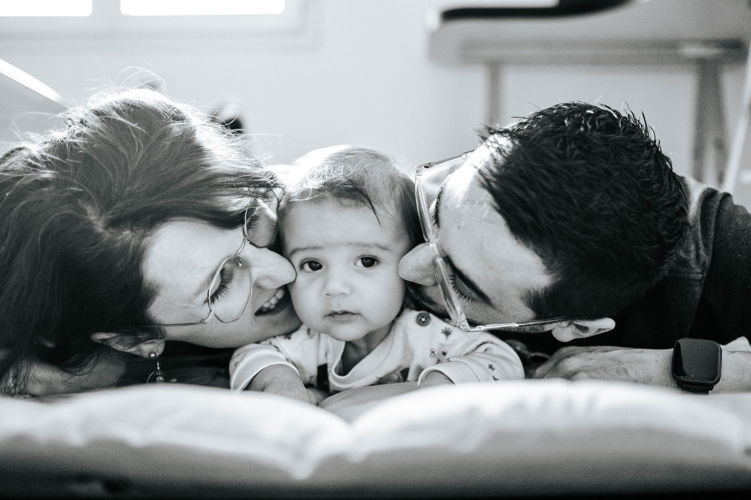 Black and white photo of two parents kissing their adorable baby on the cheeks while lying on a bed.