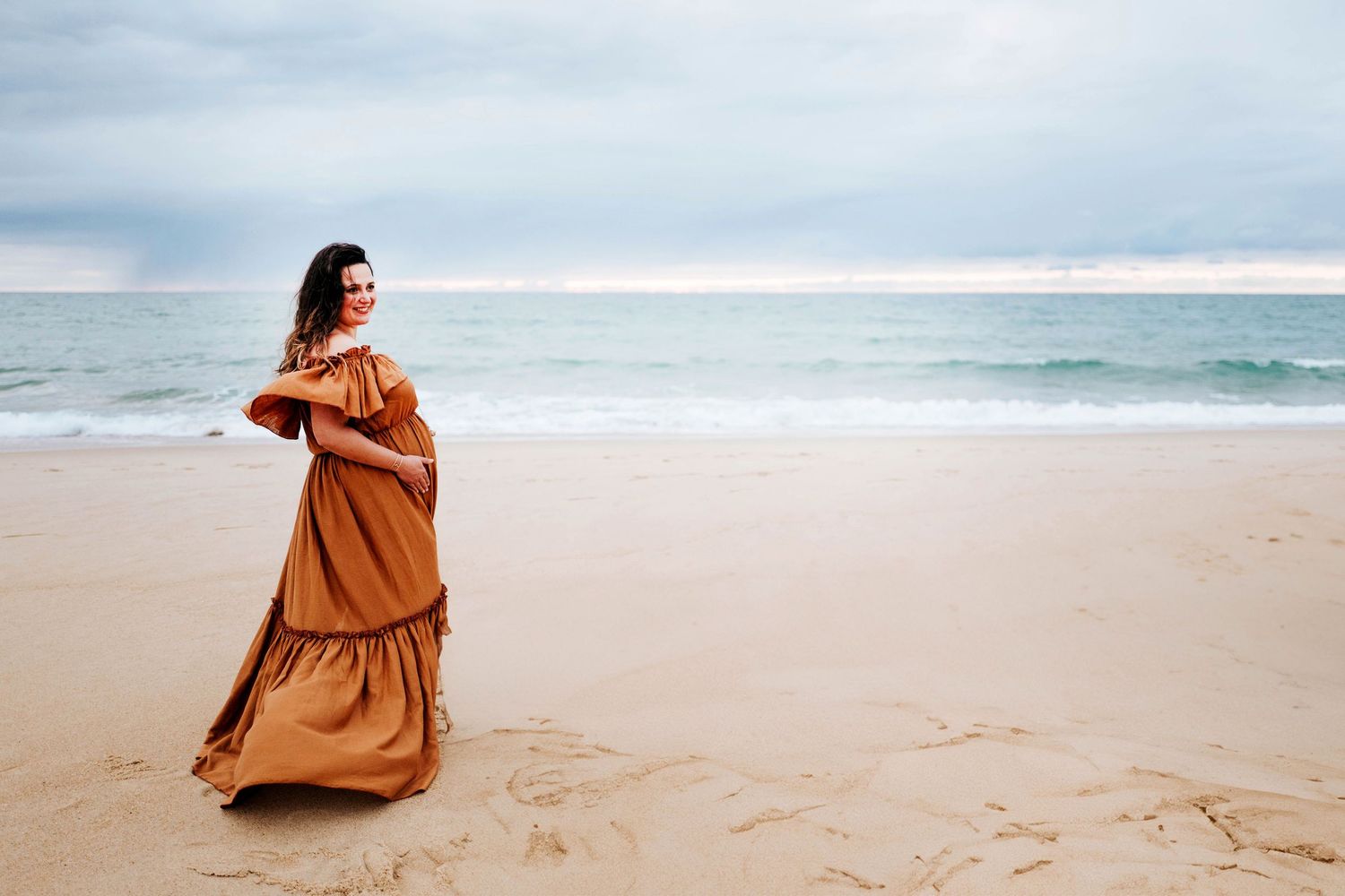 A person in a flowing rust-colored dress twirls gracefully on a sandy beach with ocean waves in the background.