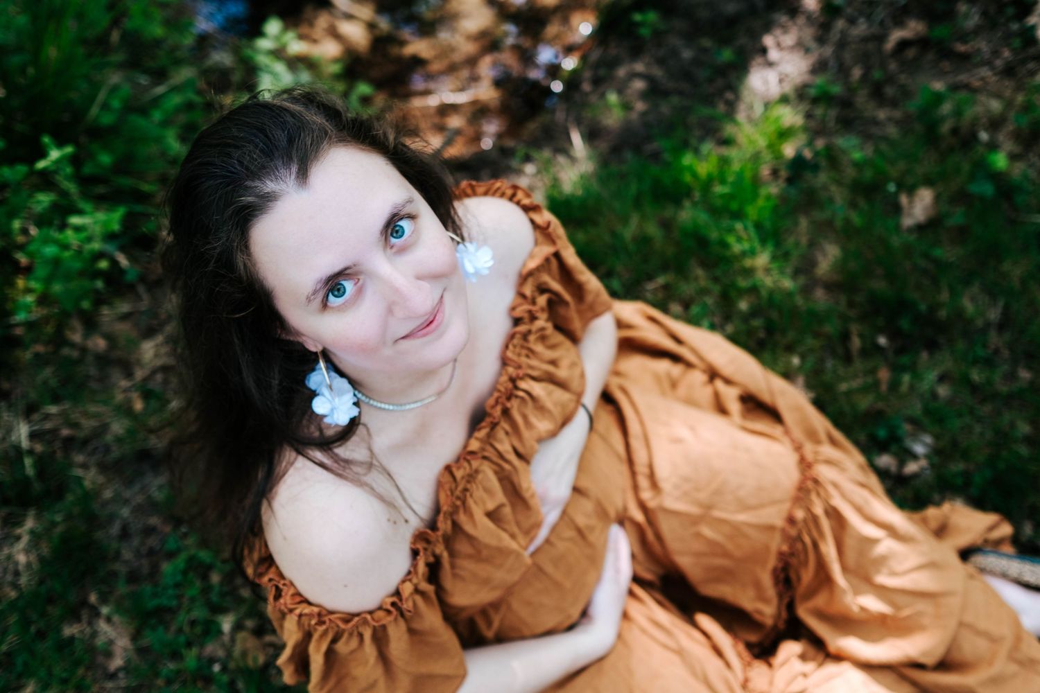Young woman in brown off-shoulder dress with white flower earrings, smiling up at camera outdoors.