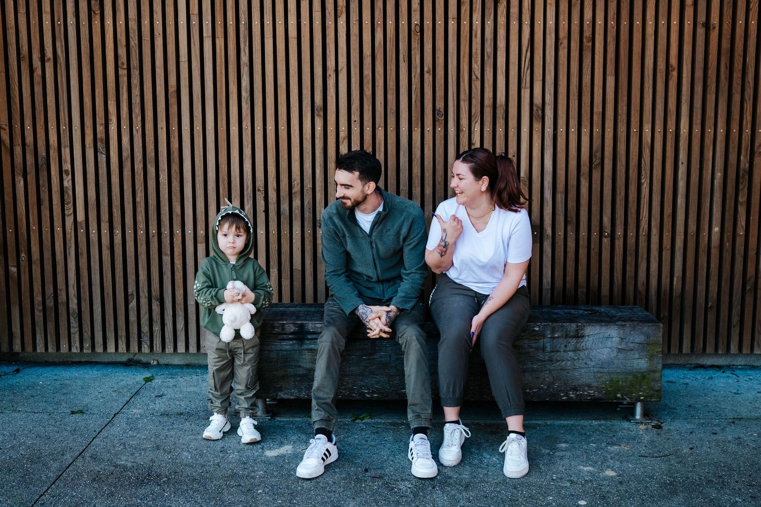 Family of three sitting on a bench against a wooden slat wall, parents smiling at each other while child holds a toy.