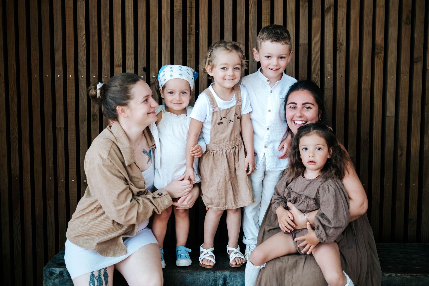 Two women and four young children pose together smiling in front of a wooden slat wall in coordinated neutral outfits.