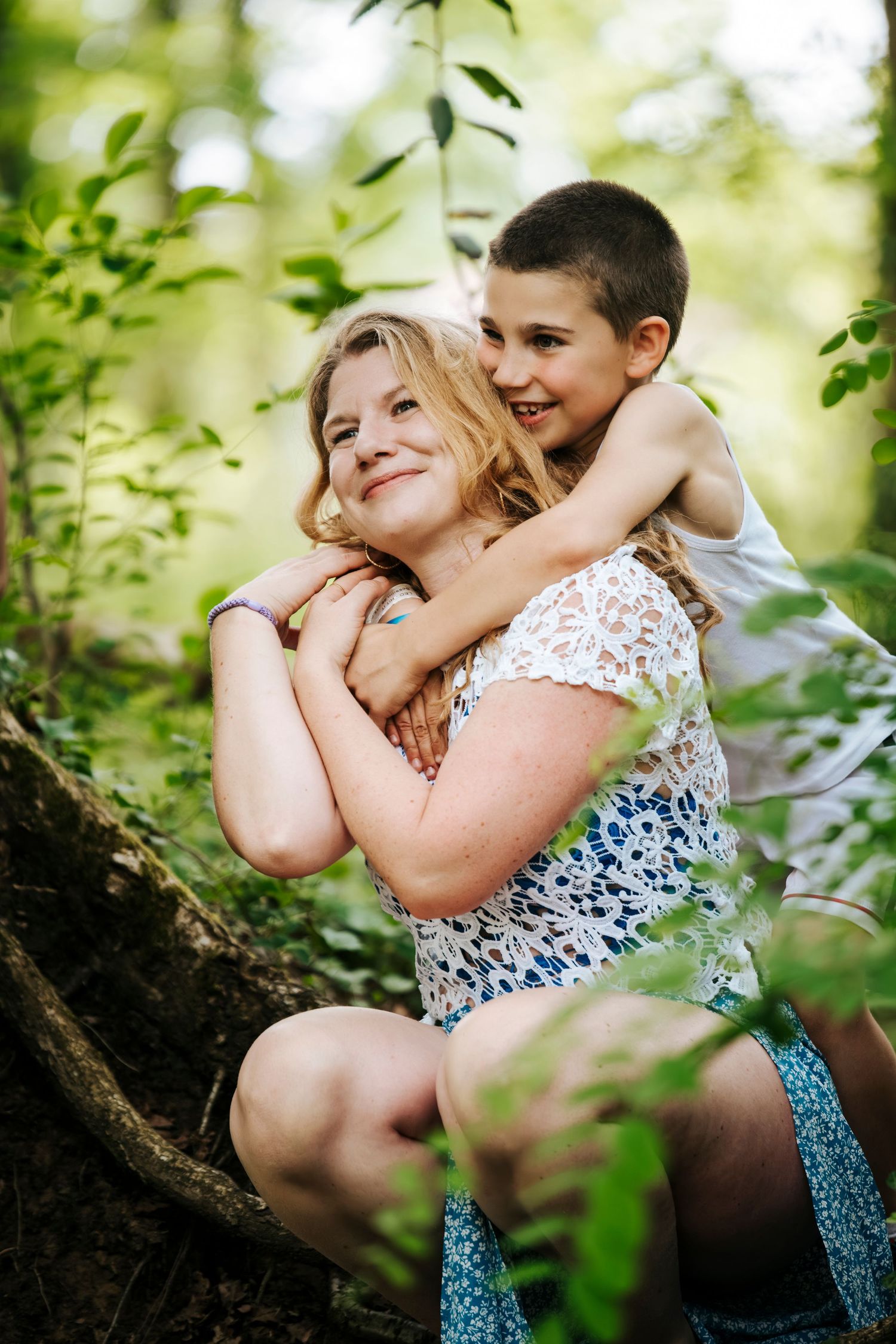 A woman and young boy share a warm hug outdoors in a lush green forest setting.