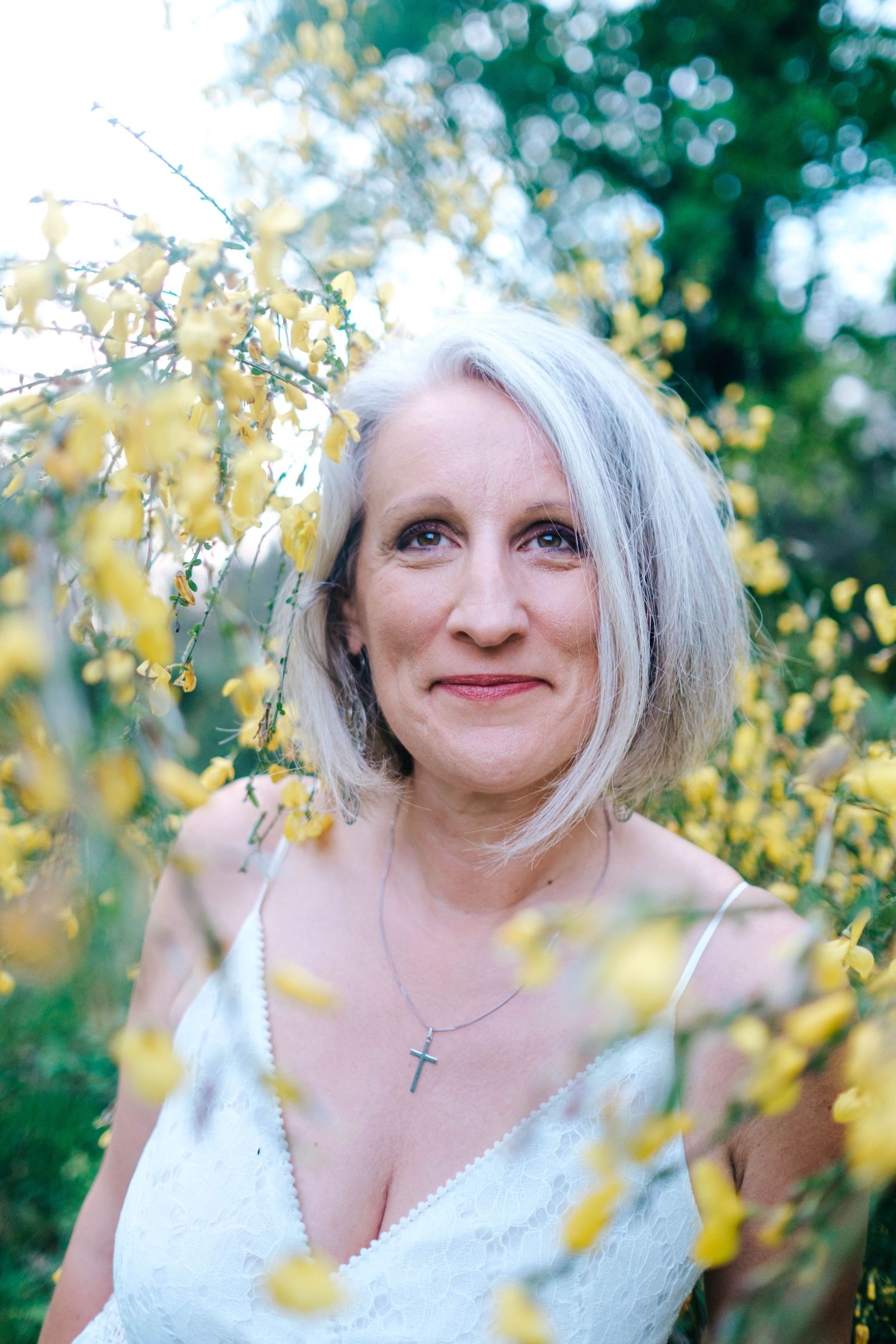 Elegant older woman with silver bob hair wearing white lace dress and cross necklace among yellow blooming flowers.