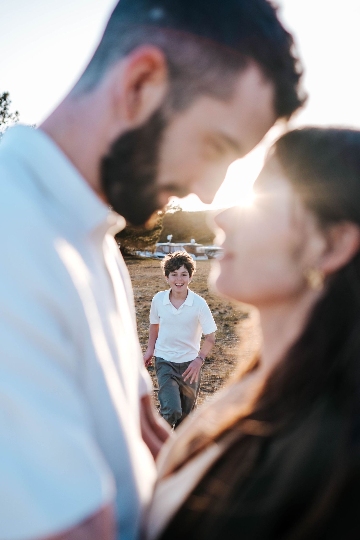 A couple shares a tender moment outdoors while a smiling child stands in the background, bathed in golden sunlight.