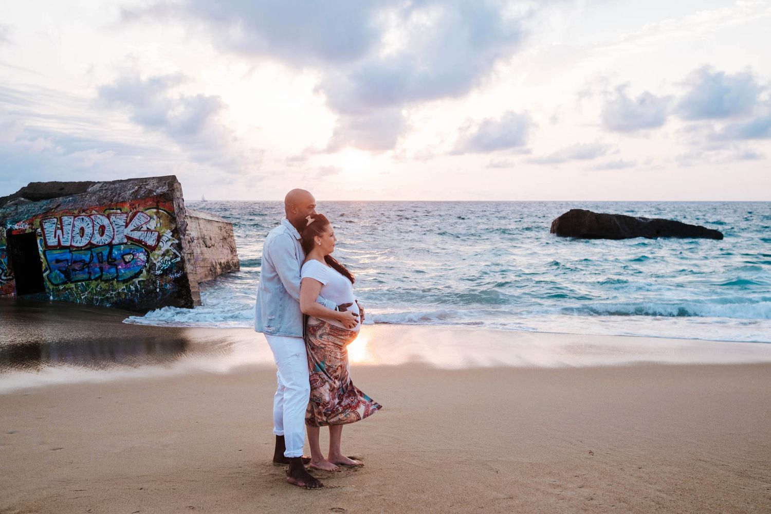 A romantic couple stands on a sandy beach at sunset with waves crashing against rocks in the background.