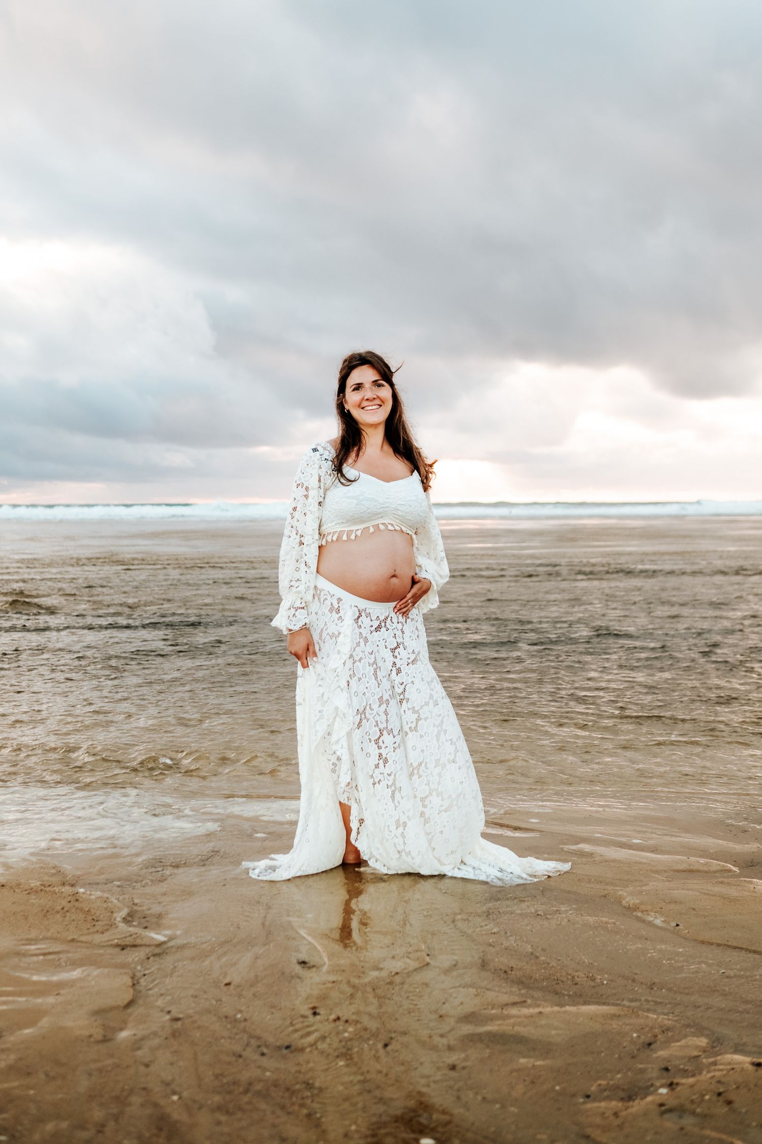 A white lace dress flows gracefully against a moody beach landscape with cloudy skies and sandy shores.