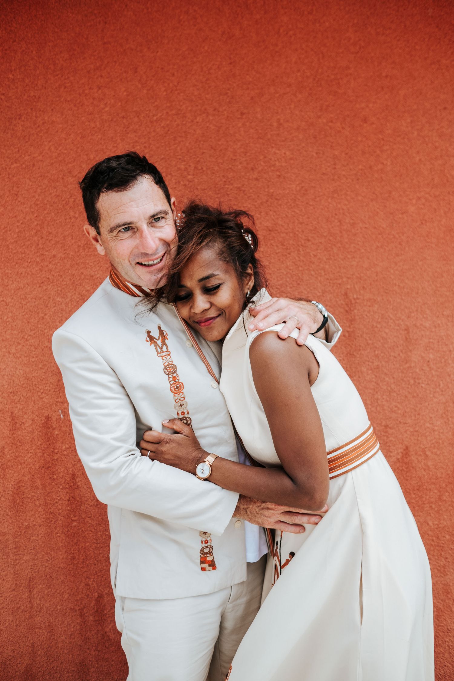 A couple in white attire embraces lovingly against a warm terracotta background.