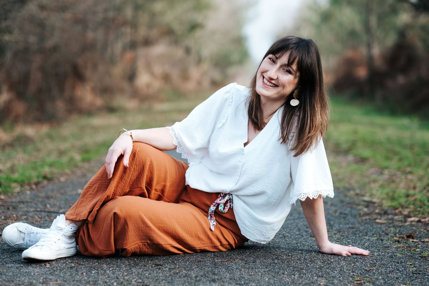 Someone in a white blouse and rust-colored wide-leg pants sits casually on the ground while smiling outdoors.
