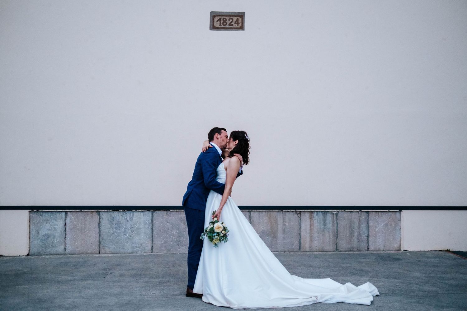 Bride and groom share a romantic kiss in front of a white wall, bride in elegant white gown holding a bouquet.
