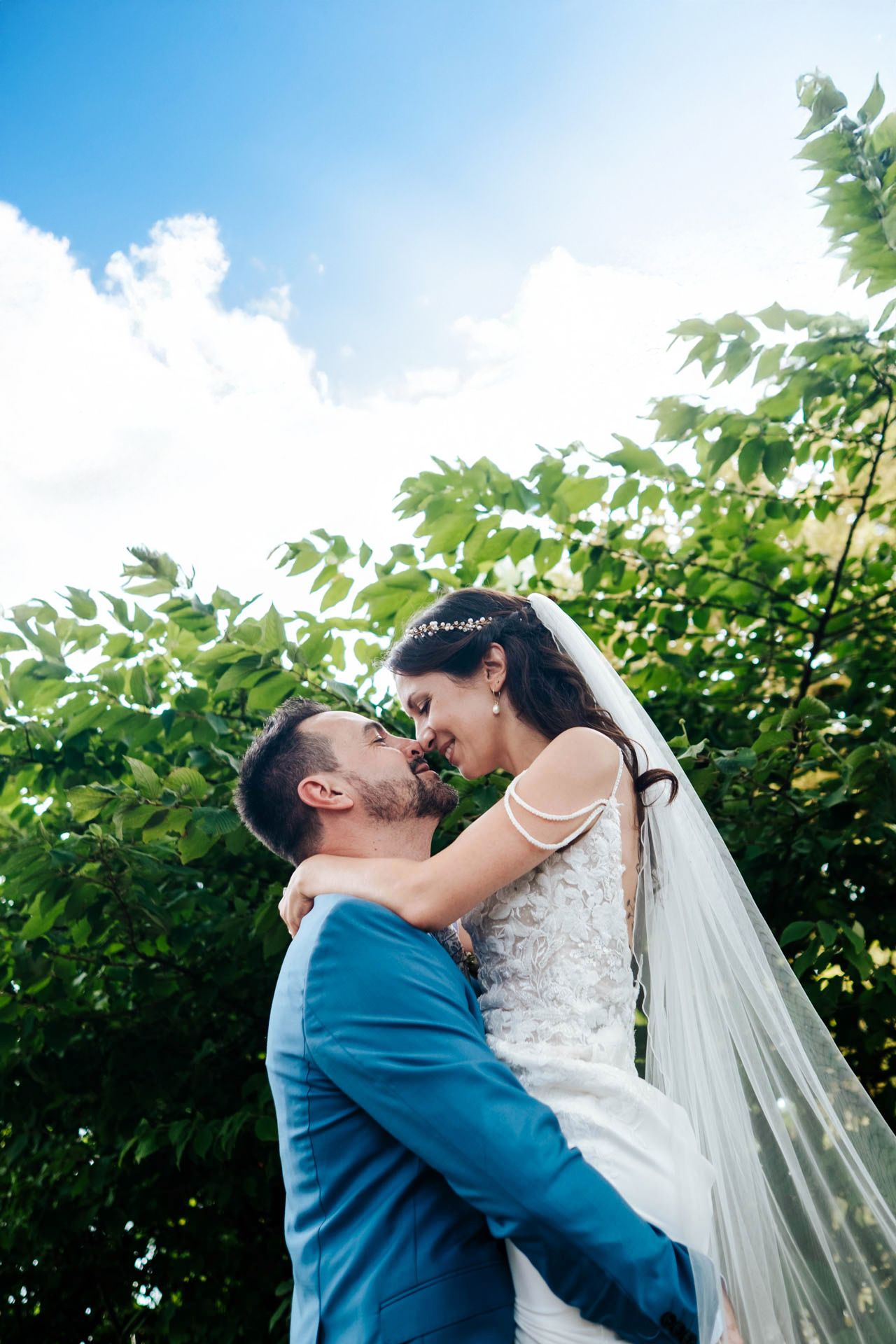 A romantic wedding moment captured outdoors with a flowing veil and blue suit against lush green foliage and blue sky.