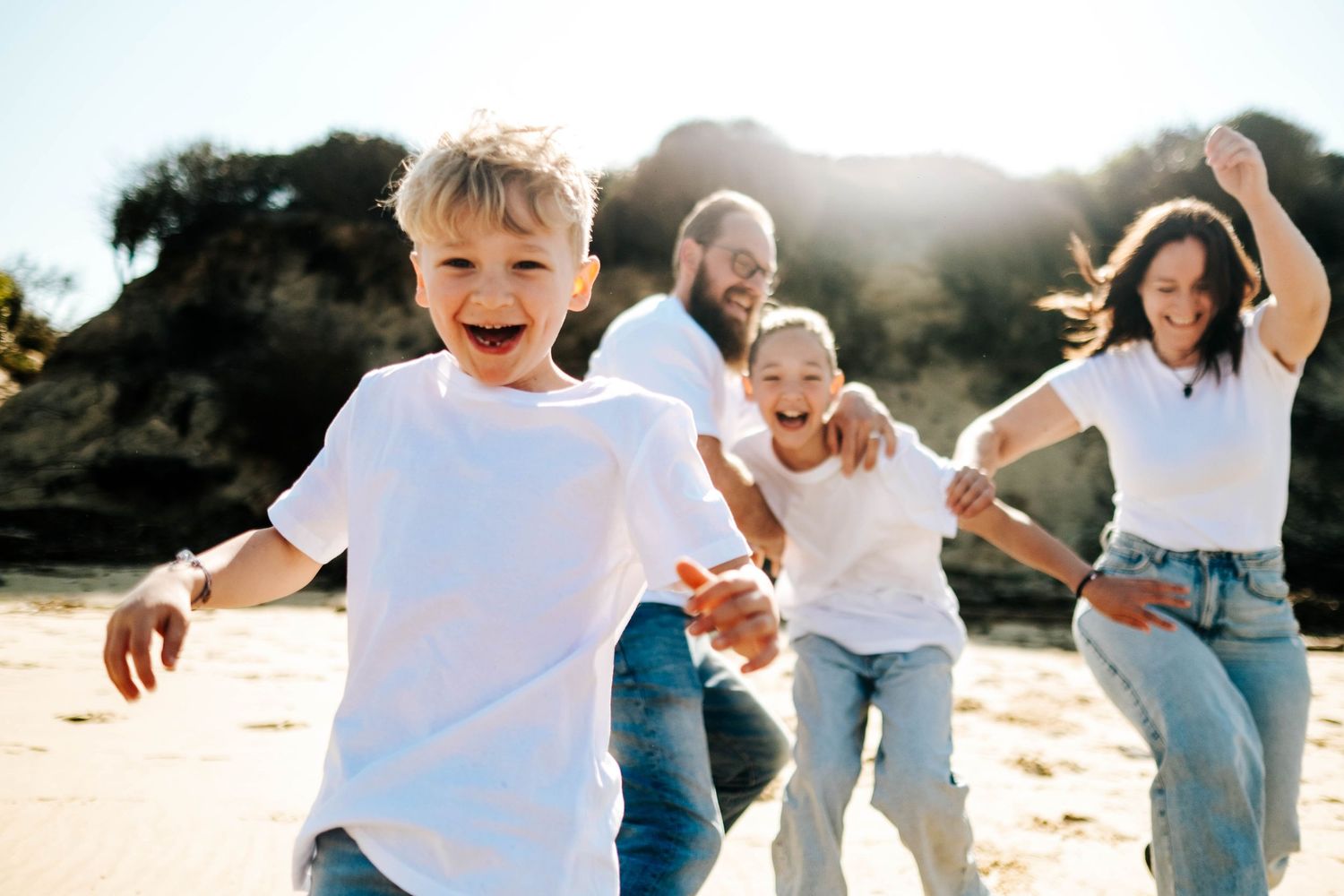 Happy family running on a sunny beach, parents and two young children laughing and playing outdoors together.