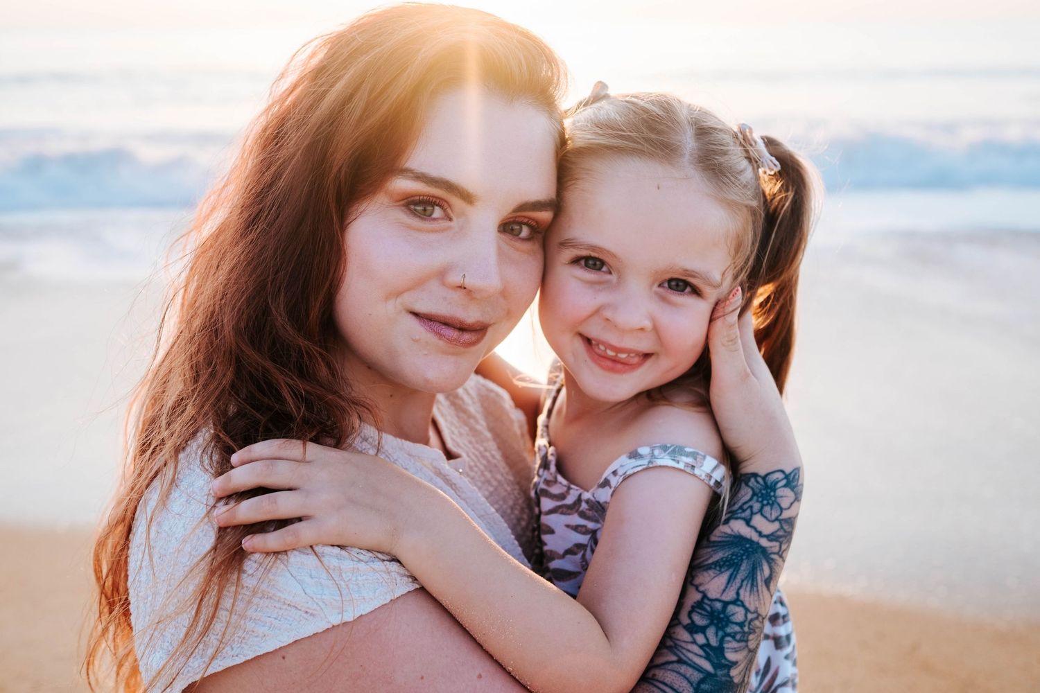 A mother and young daughter share a warm embrace on a beach at golden sunset hour.