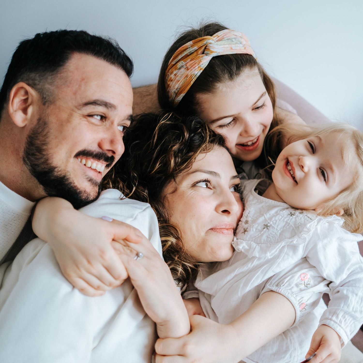 Happy family of four lying together, smiling and cuddling, with two young girls and their parents in a cozy setting.