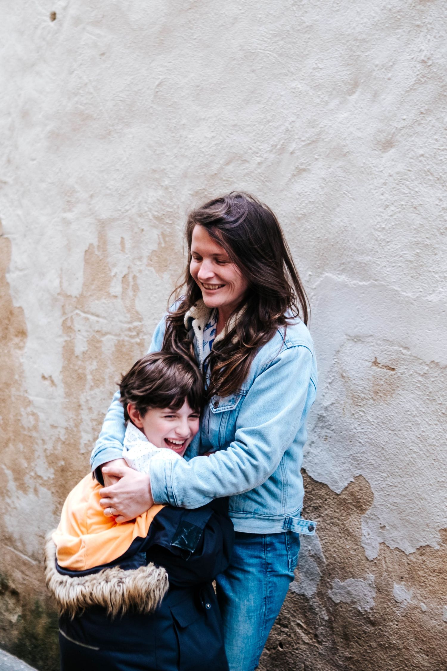 A smiling woman in a denim jacket hugs a young boy in an orange shirt against a rustic wall.
