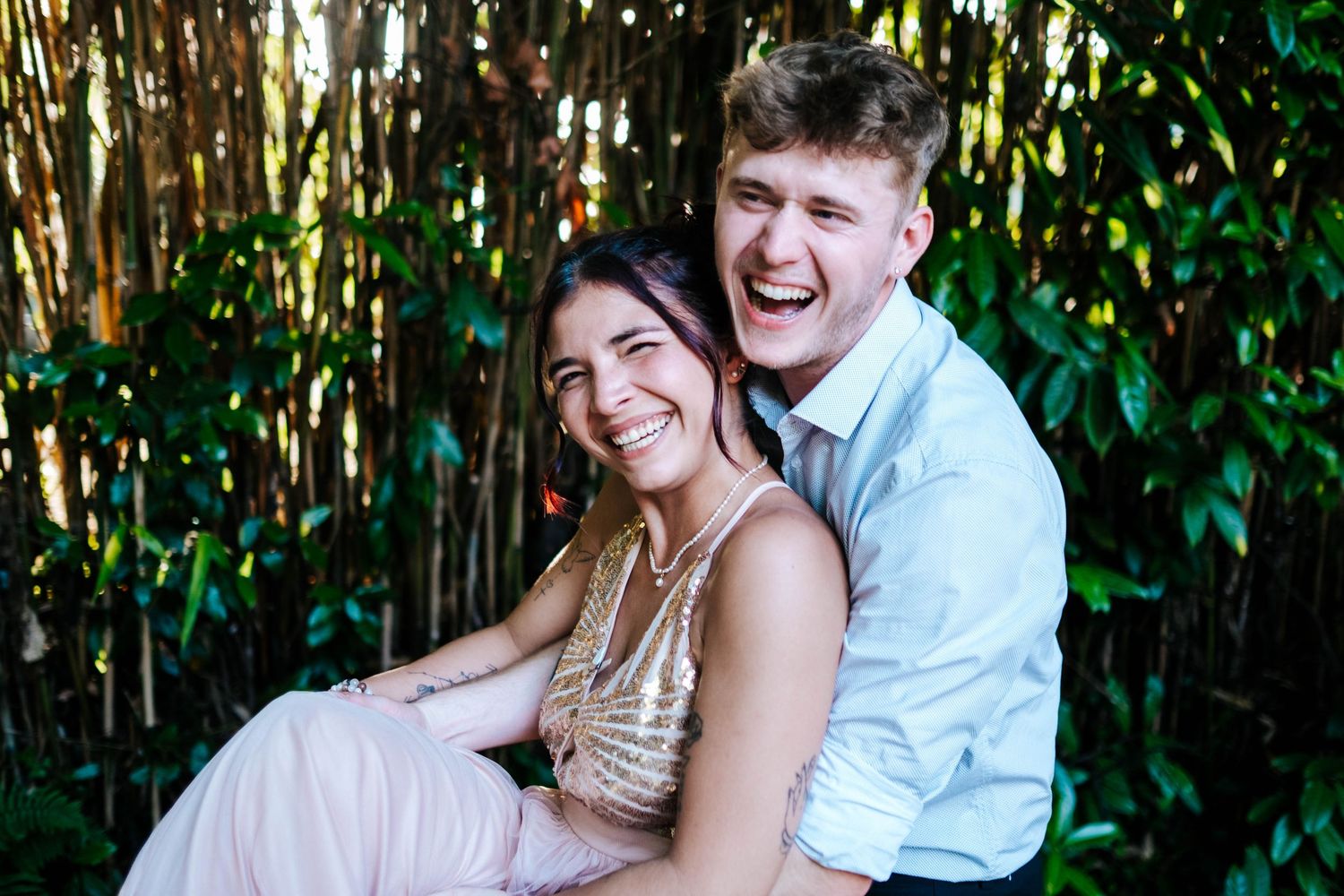 Happy young couple laughing together outdoors, wearing formal attire in front of lush green bamboo backdrop.
