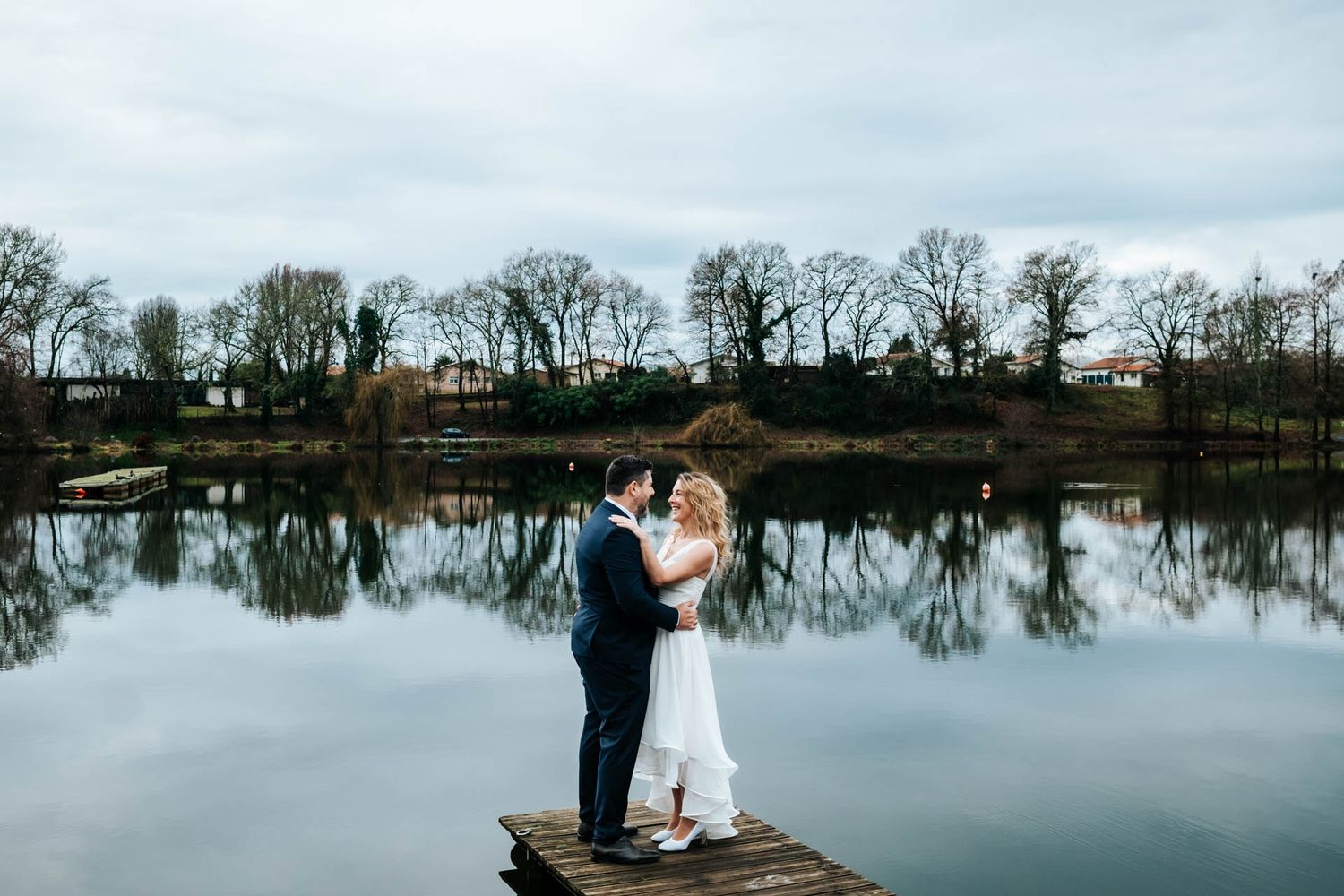 A romantic photoshoot by a serene lake with trees reflecting in the still water.