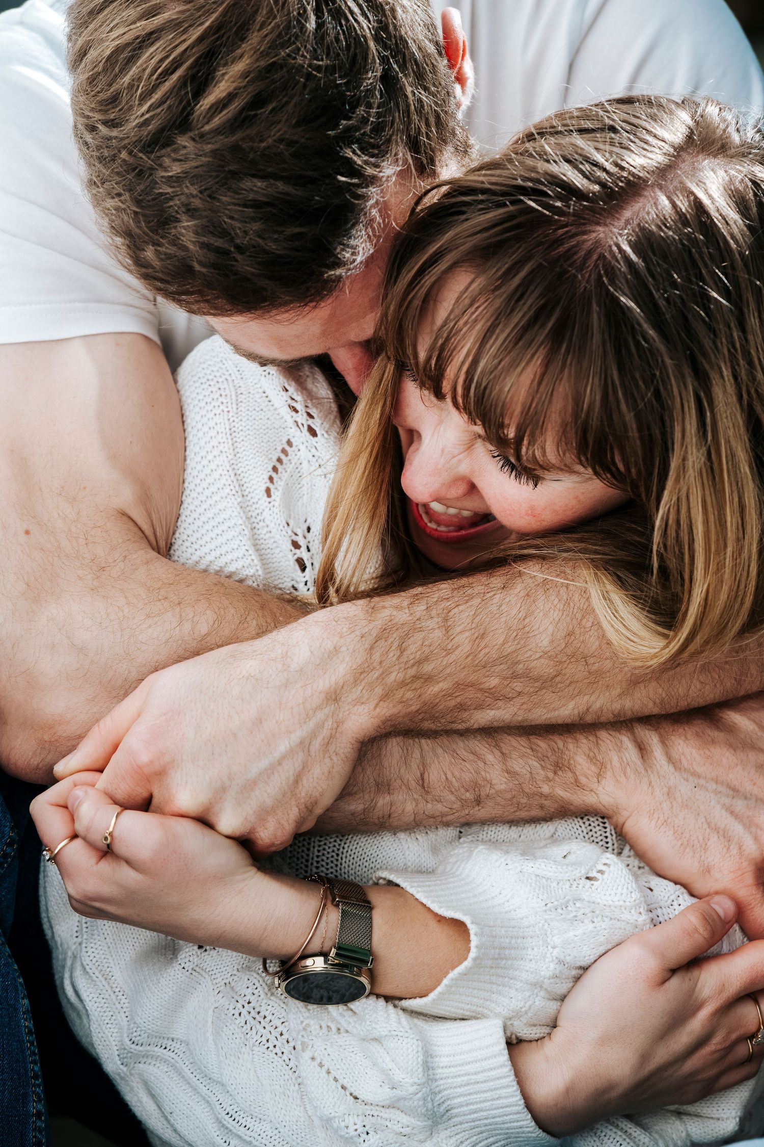 A couple sharing a tender embrace, the man kissing the woman's head while holding hands, both smiling warmly.