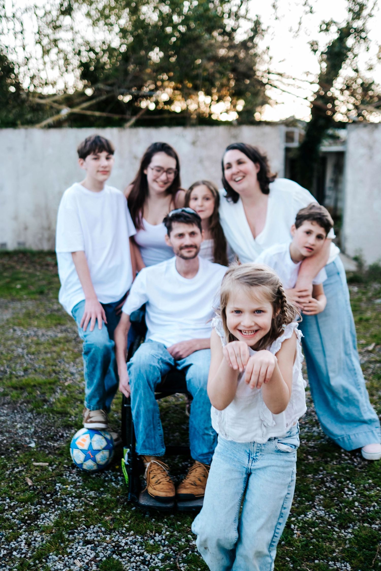 A joyful family of seven poses outdoors, with a young girl in front smiling, parents and children behind her.