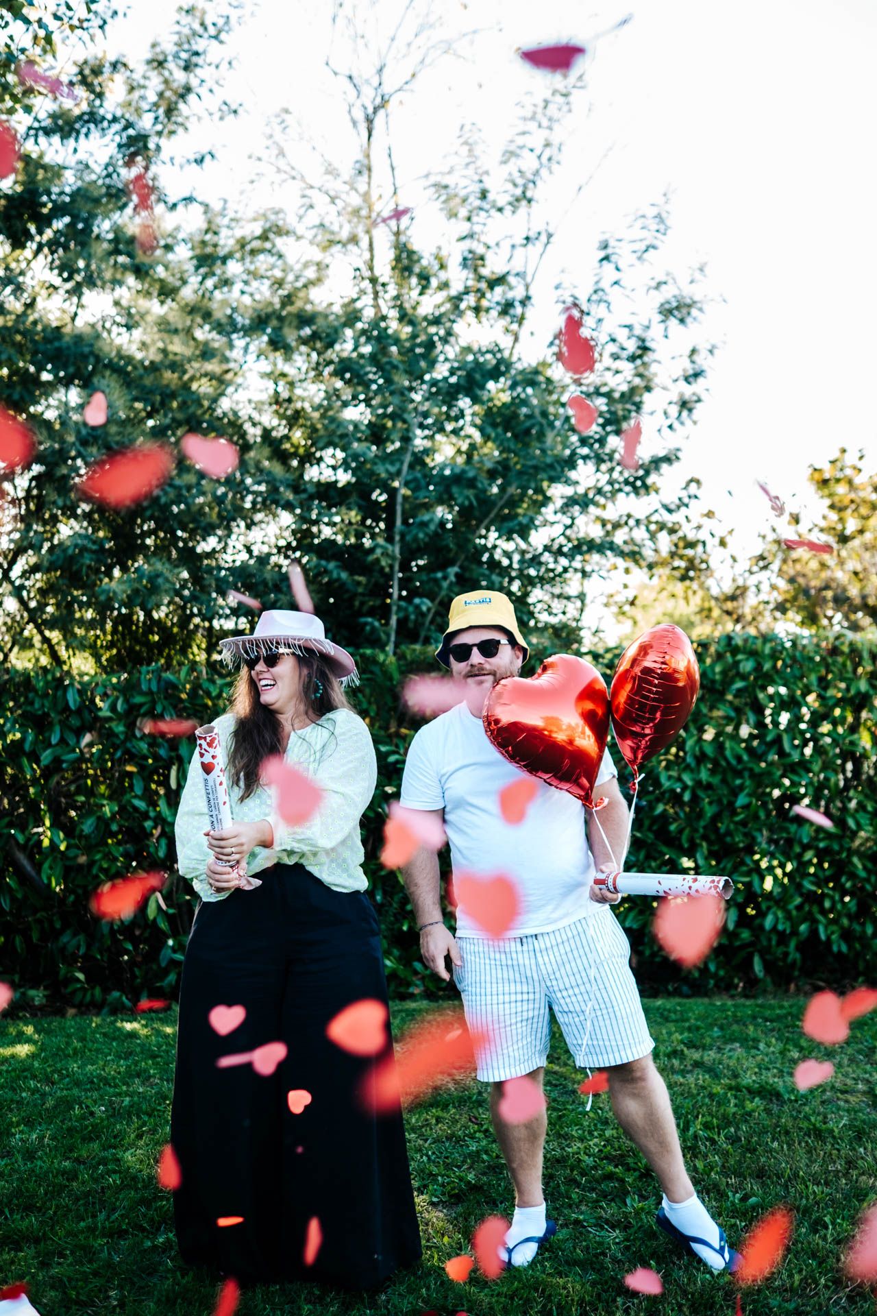 Couple in hats and sunglasses celebrating outdoors with red heart confetti and balloon in a lush green garden.