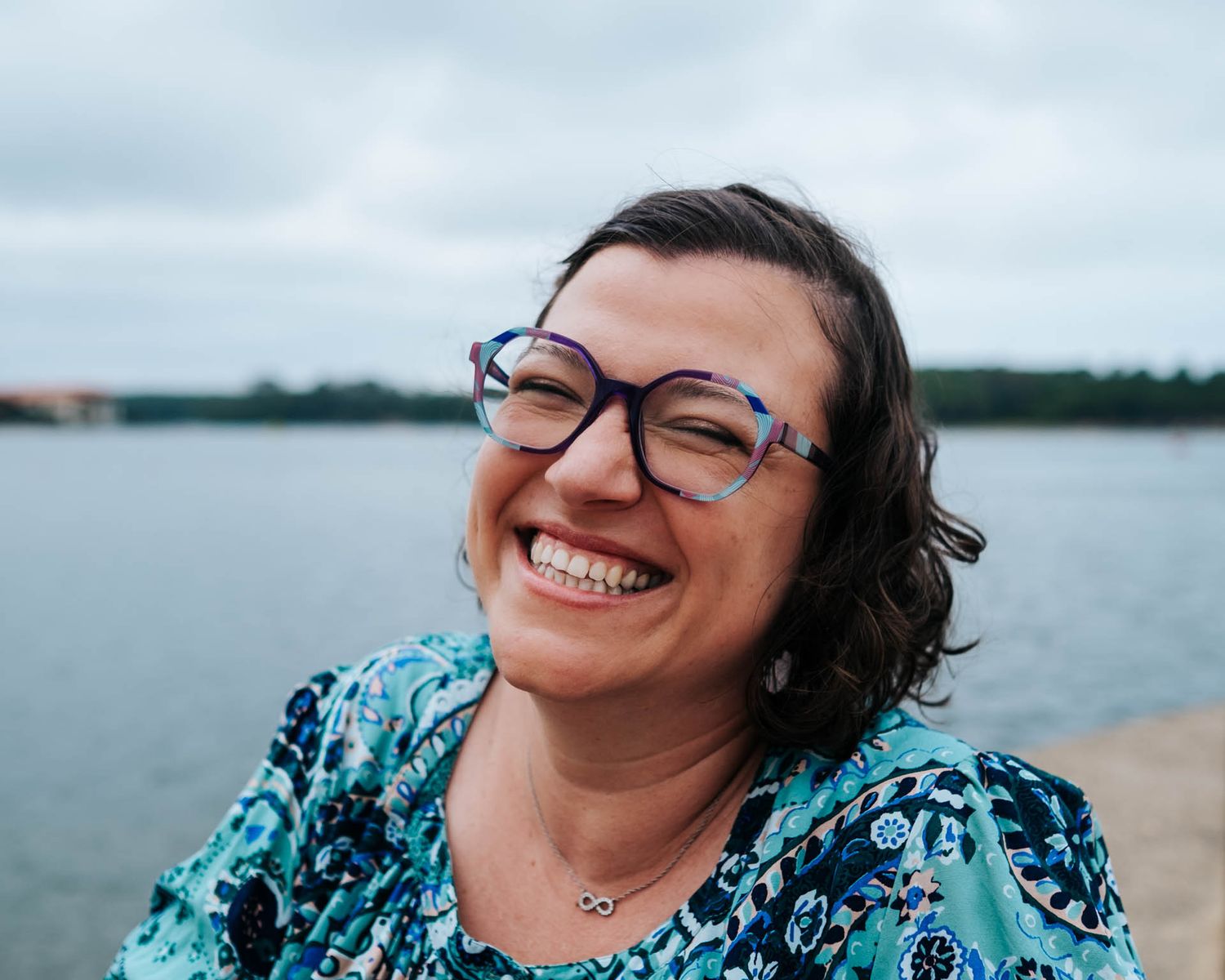 Someone with glasses and a bright smile wearing a blue patterned top sits by a lake on a cloudy day.