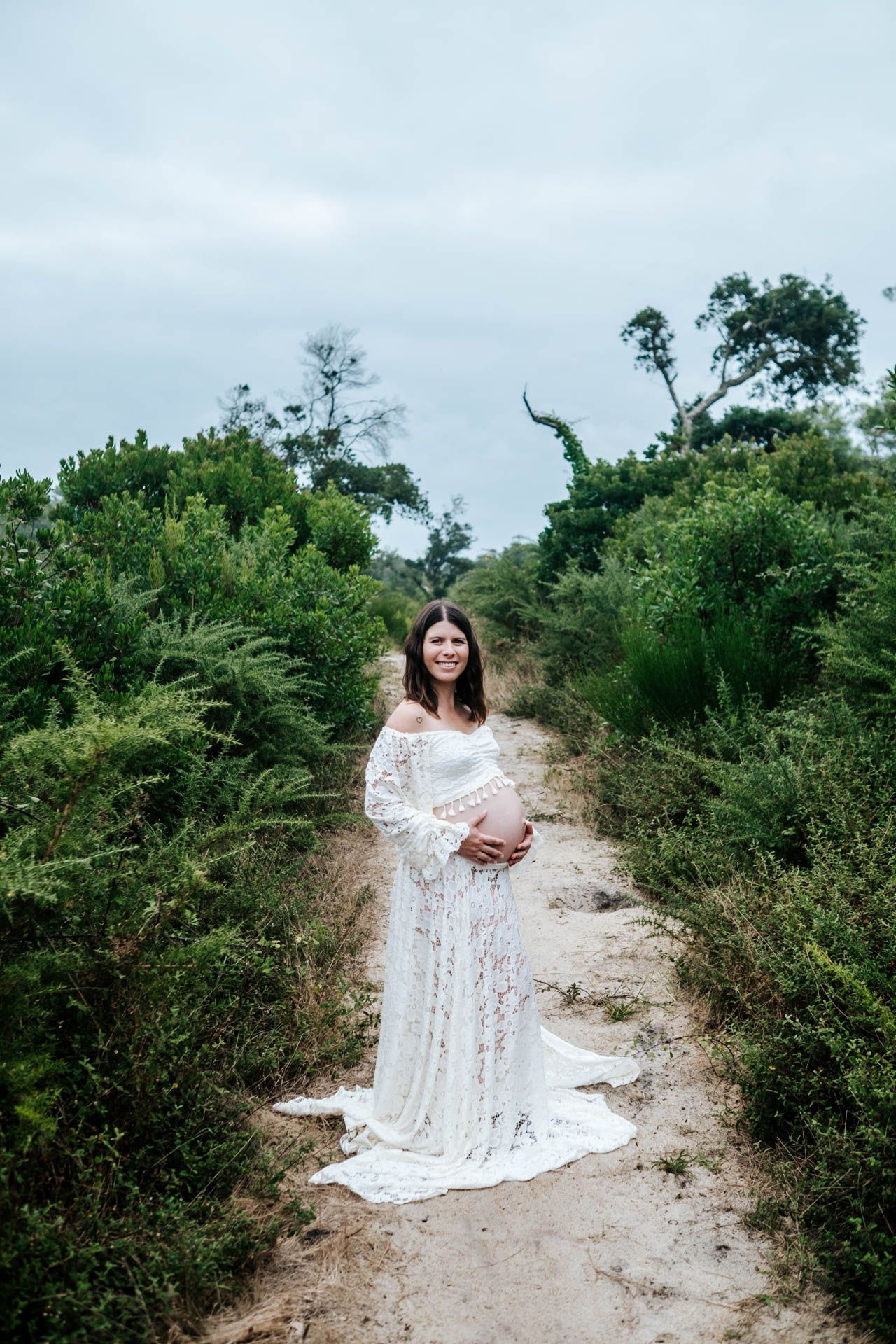 Two figures in flowing white lace dresses pose together on a sandy path surrounded by green coastal vegetation.