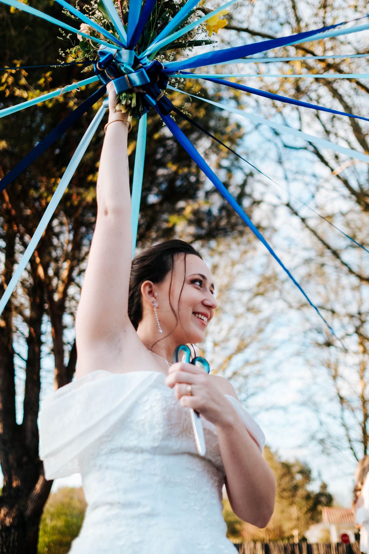 A bride in a white dress holds a blue and turquoise umbrella outdoors on a sunny day.
