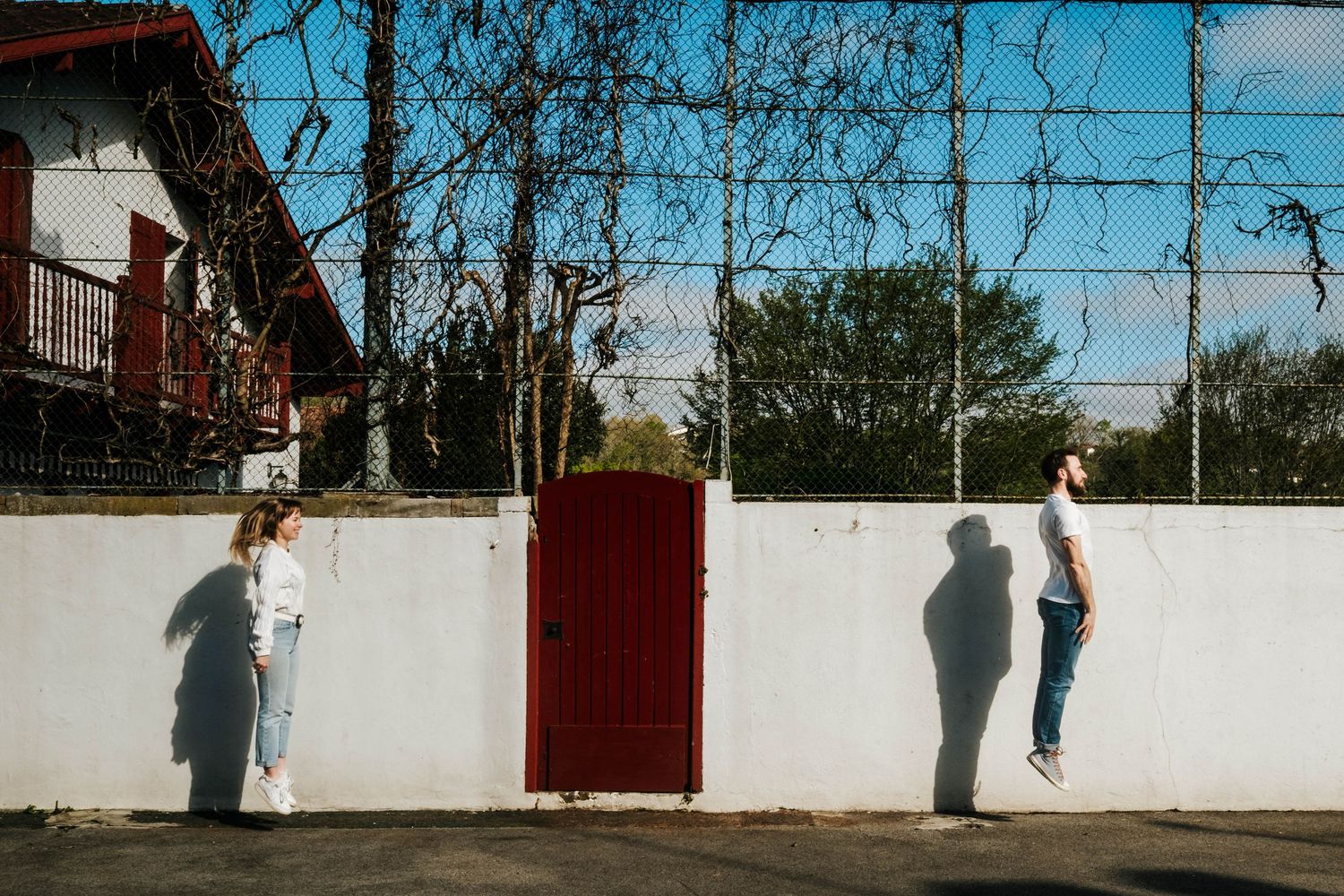 Two people stand apart against a white wall with a red gate, casting shadows in bright sunlight near a chain-link fence.