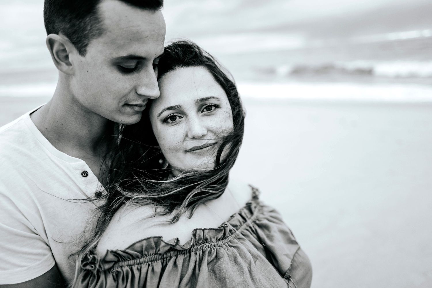 A romantic black and white portrait of a couple embracing outdoors with wind-blown hair.