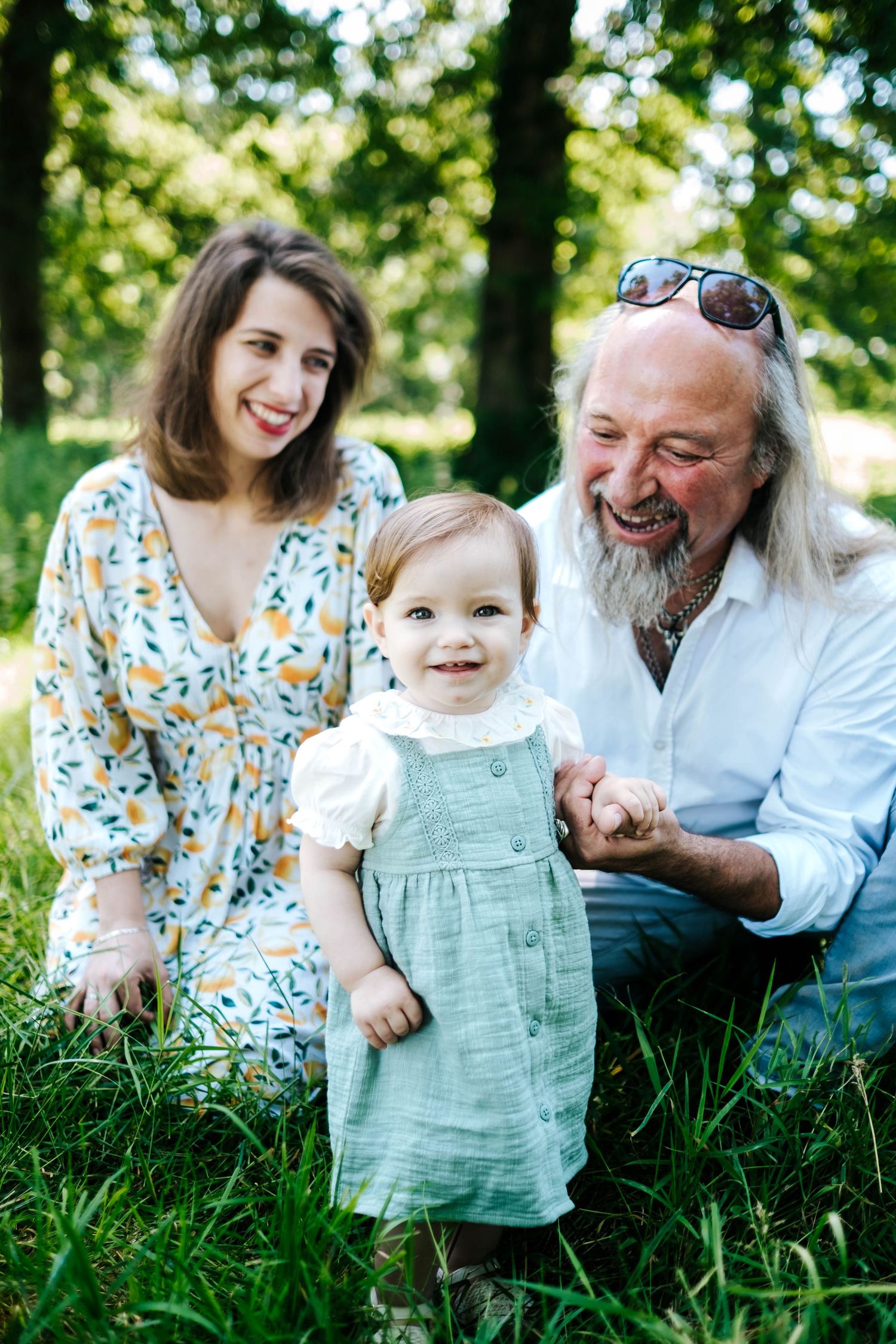 A smiling family with a young toddler girl in a green dress posing outdoors in a lush green park.