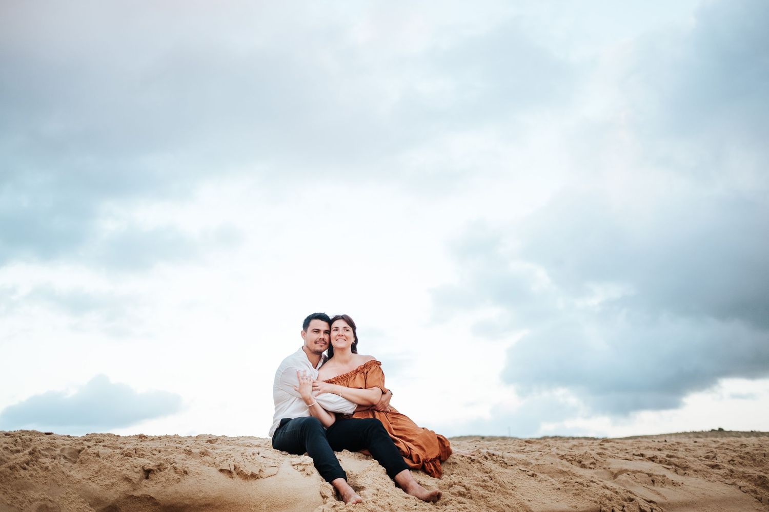 A couple sits close together on a sandy dune beneath dramatic cloudy skies.