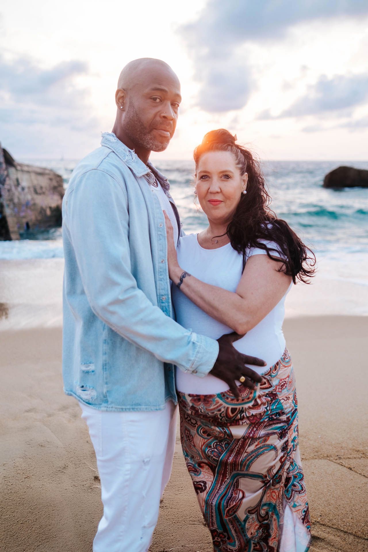 A couple embraces on a sandy beach at sunset with ocean waves and cliffs in the background.