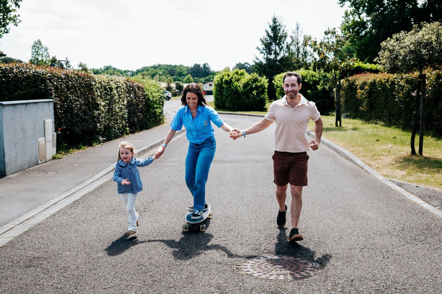 Family of three holding hands on a sunny road, mother skateboarding with young daughter and father smiling outdoors.