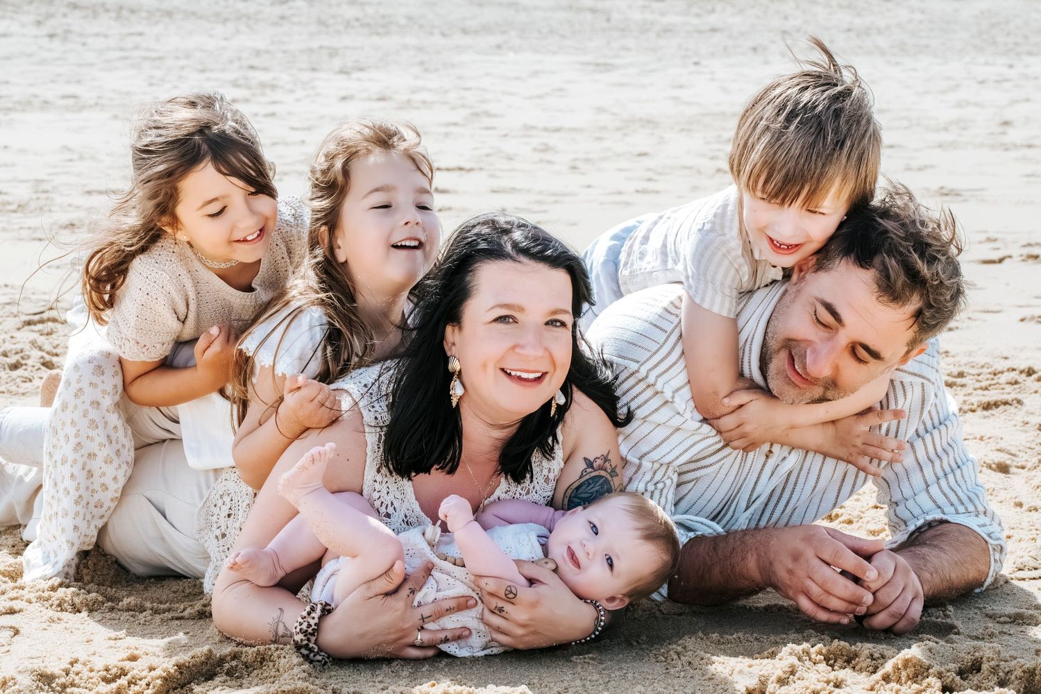 Happy family with three kids and a baby lying on sandy beach, smiling and laughing together on a sunny day.