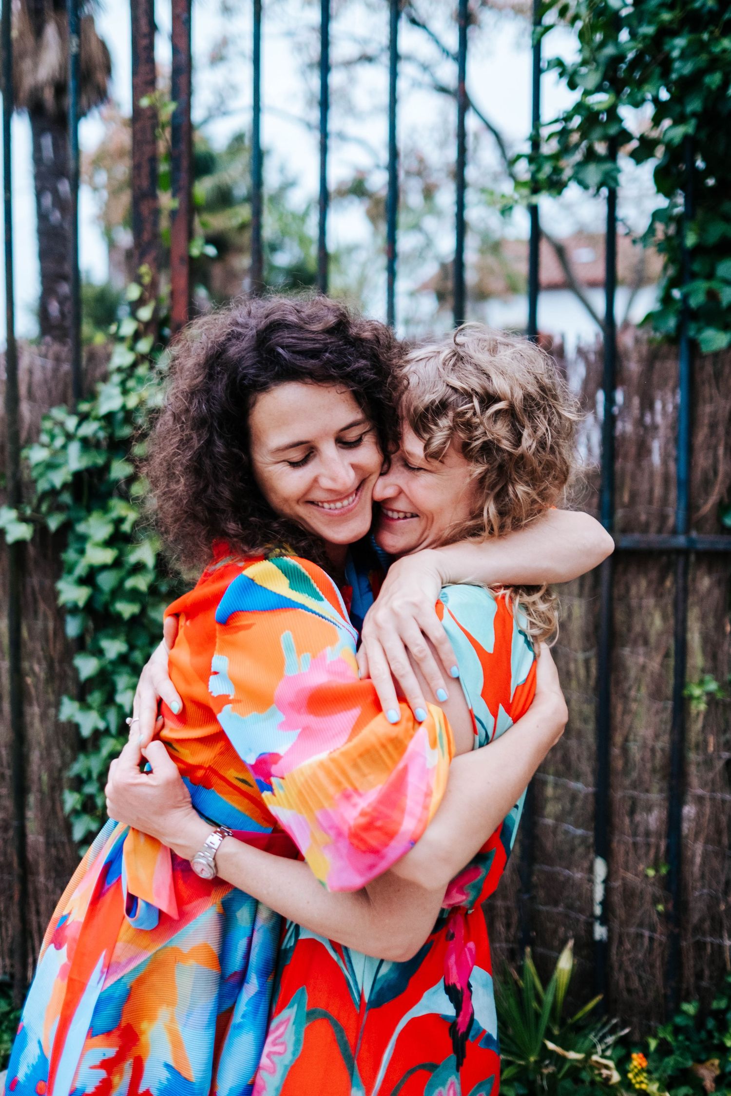 Two women in colorful floral dresses share a warm embrace outdoors near a wooden fence with green ivy.