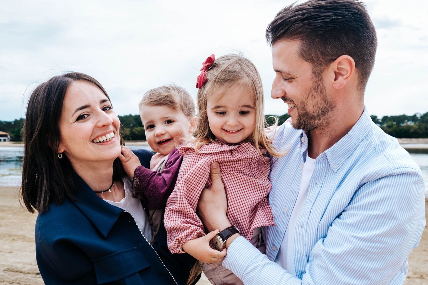Happy family with two young girls smiling outdoors near a beach, parents holding children in a warm embrace.