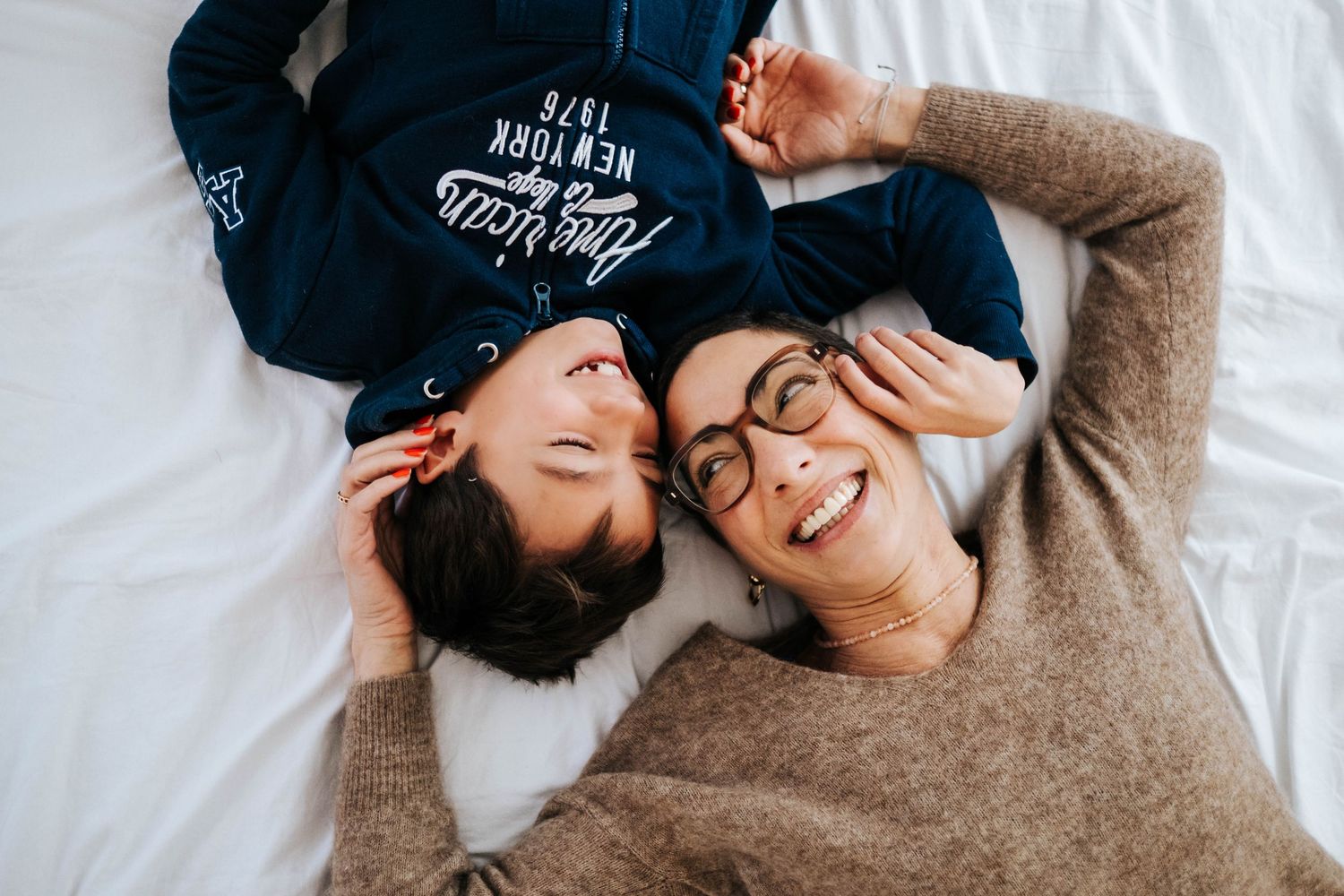 Two women lying on a white bed, smiling and laughing together in a joyful candid moment.