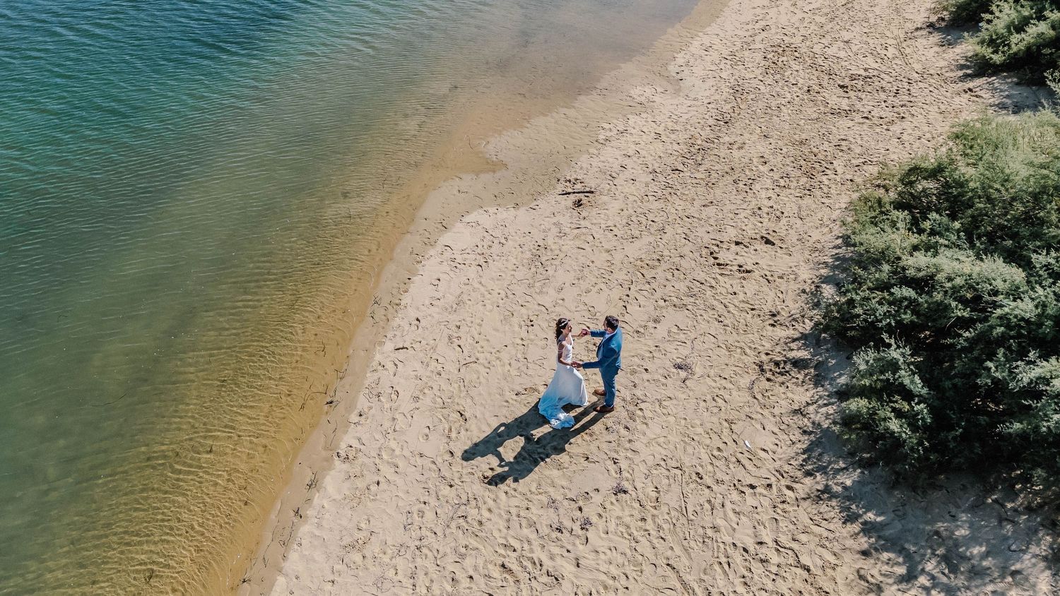 Aerial view of a couple standing on a sandy beach shore beside calm blue water surrounded by trees.