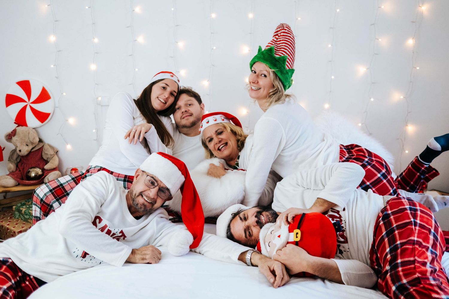 Family members in matching red plaid pajamas lying on white bedding with holiday lights in the background.