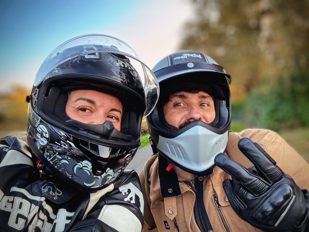 Two motorcycle riders wearing protective helmets and gear pose for a selfie outdoors on a sunny day.