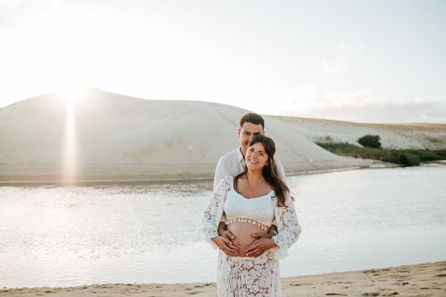 Couple embraces on sandy beach during sunset with mountains in background.