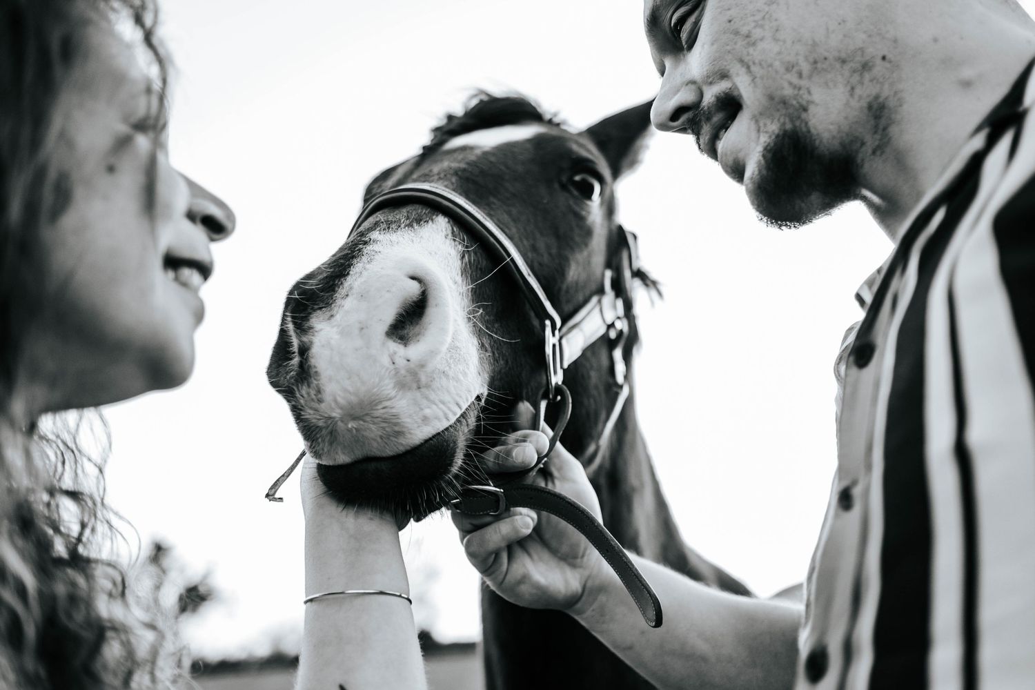 Black and white photo of a young couple bonding with a horse outdoors, gently holding its halter.
